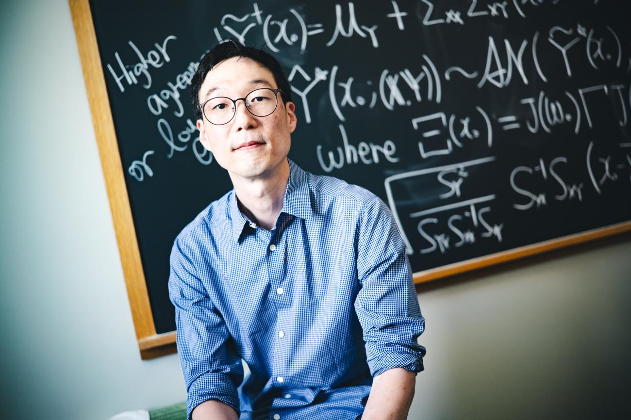 Taeho Kim wearing a blue button-up shirt and glass sitting in front of a black board with math problems on it.